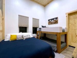 Cabin bedroom in Red River Gorge rental with a queen bed featuring blue bedding, a yellow pillow, and a decorative pillow reading "Our Happy Place." Bunk beds and mountain-themed artwork add to the cozy atmosphere.