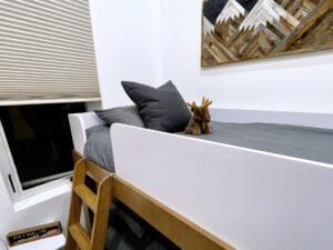 A cozy bunk bed in a cabin bedroom, featuring gray bedding, a cute moose plush, and a wood-crafted mountain art piece. A window with blinds offers natural light, enhancing the rustic charm, perfect for a Red River Gorge rental cabin.