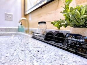 Granite bathroom countertop in a Red River Gorge cabin rental. A wire basket holds makeup remover wipes and dark washcloths, next to a mason jar of cotton swabs. A green plant adds a touch of nature.