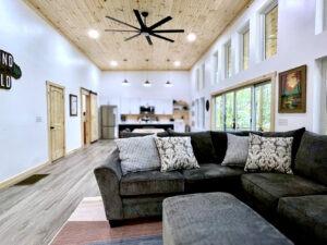 Cabin living room in Red River Gorge, Kentucky. A comfy gray sectional faces a modern kitchen. Wood ceiling and large windows create a spacious feel. Perfect for a relaxing cabin stay.