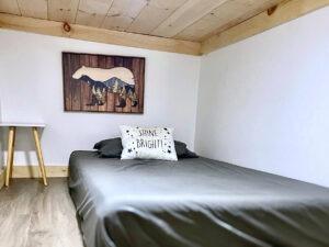 Cozy cabin bedroom in Red River Gorge rental, featuring a twin bed with gray sheets, a "Shine Bright!" pillow, and a wooden bear art piece. Wood ceiling and flooring add to the rustic charm.