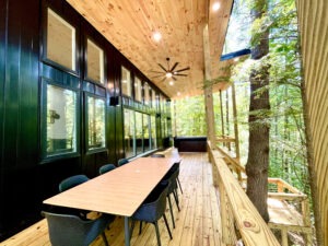 Deck of a Red River Gorge rental cabin with a long dining table and chairs, overlooking a lush forest. A hot tub sits at the far end. The cabin features black siding and a wood ceiling.