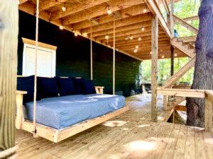Relax on a wooden porch swing at a Red River Gorge rental cabin. The swing, with dark cushions, hangs under string lights. Stairs lead up to the cabin, nestled among trees.