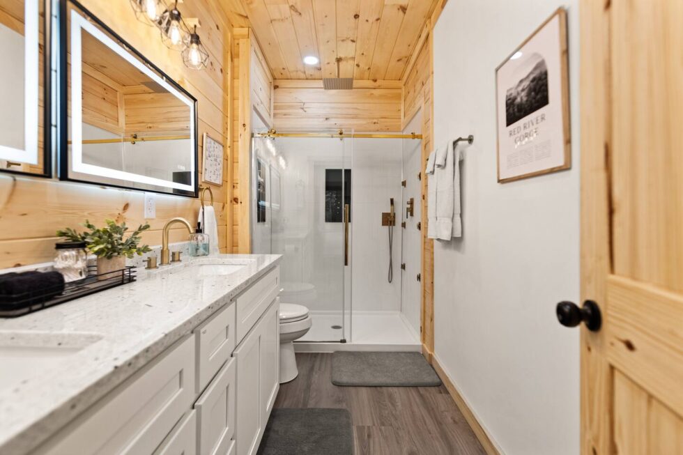 Bathroom in a Red River Gorge rental cabin. Features wood paneled walls, a white vanity with speckled countertop, a glass-enclosed shower, and a framed "Red River Gorge" print.