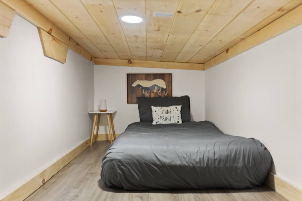 Cozy sleeping nook in a Red River Gorge rental cabin. A gray bed with a "Shine Bright!" pillow sits under a natural wood ceiling with a skylight, next to a small bedside table with a lamp.