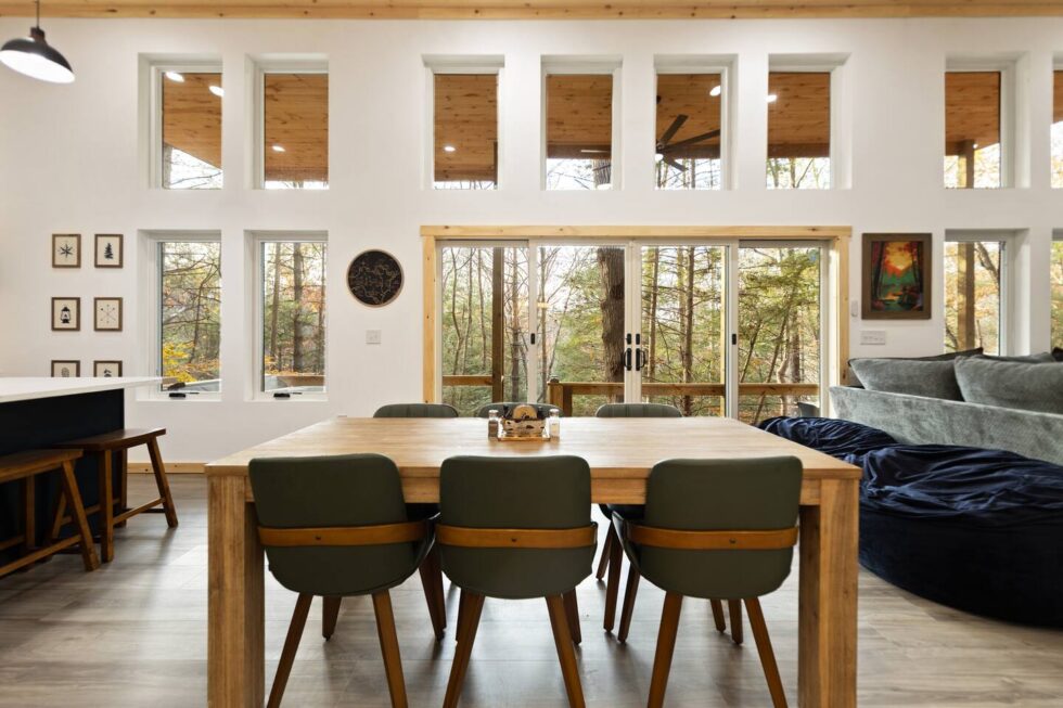 Cabin dining area with a large wood table, green chairs, and expansive windows showcasing the Red River Gorge forest. Natural light fills the room, highlighting the open space and rustic-modern design of this Kentucky rental cabin.
