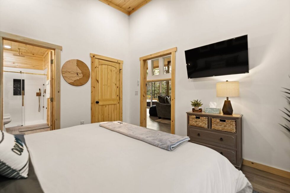 Cozy Red River Gorge cabin bedroom with a comfy bed, TV, dresser, and views into the bathroom and living area. Wood trim and high ceilings add to the rustic charm.