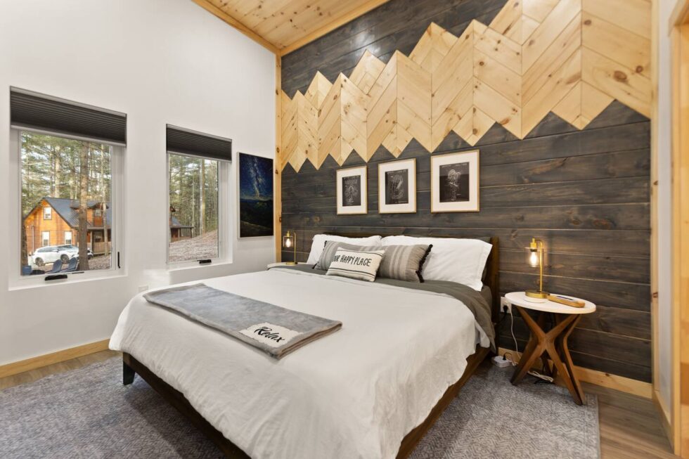 Cozy Red River Gorge cabin bedroom with a king bed, white linens, and a decorative wood accent wall. Windows offer views of the Kentucky forest outside. "Relax" throw blanket on the bed.
