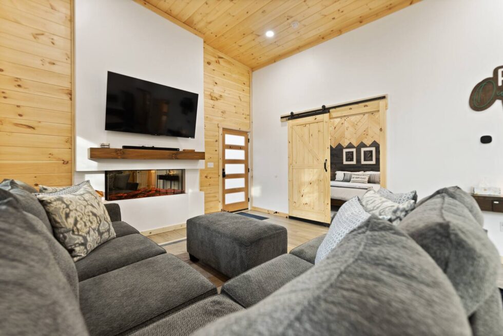 Cozy Red River Gorge cabin living room with a gray sectional sofa, modern fireplace, and barn door leading to a bedroom. Wood paneled ceiling and walls add warmth to this Kentucky rental cabin.