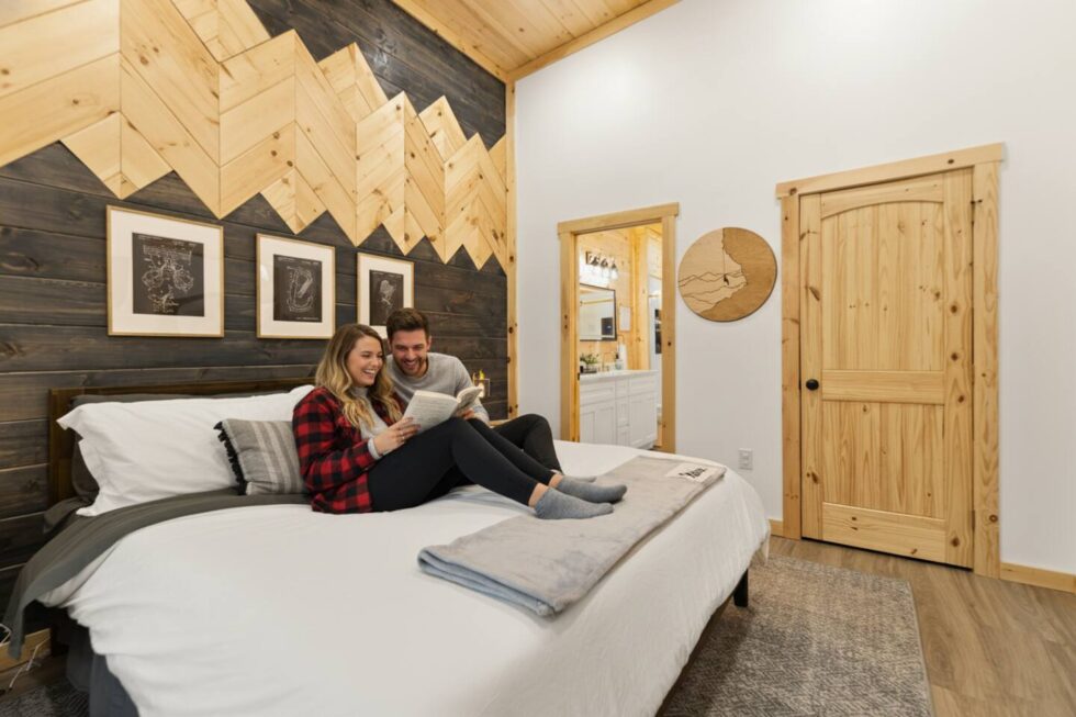 A couple relaxes on a bed in a Trickle Creek Retreat cabin in Red River Gorge, KY. The room features a wood accent wall, white bedding, and a cozy, rustic feel, perfect for a relaxing getaway.