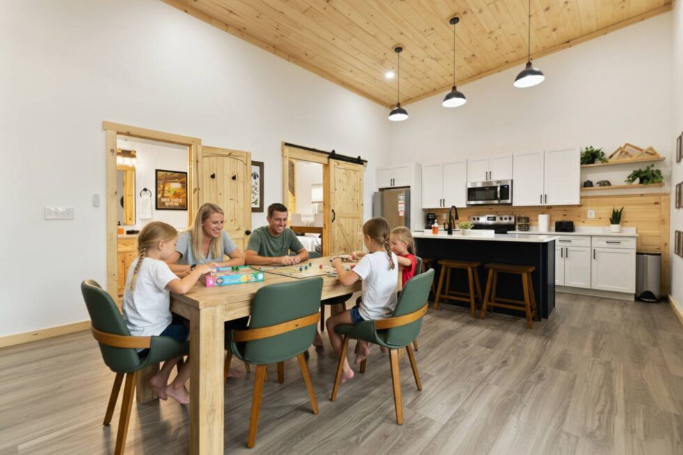 Family plays a board game at the dining table of a Red River Gorge cabin rental. The modern cabin features a wood ceiling, white walls, and a kitchen with a black island. A sliding barn door leads to a bedroom.
