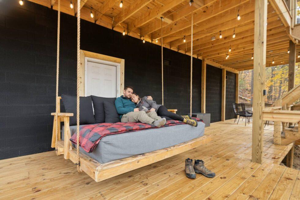 Couple relaxing on a porch swing at a cozy rental cabin in Red River Gorge, Kentucky. String lights illuminate the wooden deck, creating a warm, inviting atmosphere.