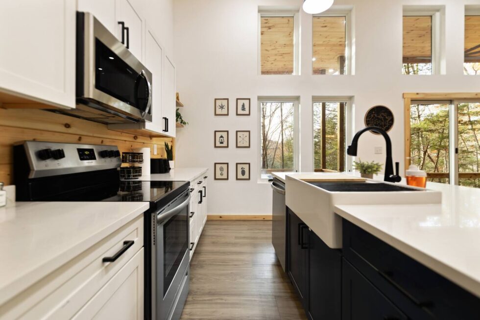Modern cabin kitchen in Red River Gorge rental with white cabinets, black hardware, stainless steel appliances, and a farmhouse sink. Large windows offer views of the Kentucky forest.