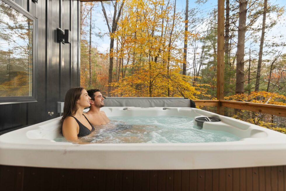 A couple relaxes in a hot tub at Trickle Creek Retreat, a cabin rental in Red River Gorge, KY. Fall foliage surrounds the deck, offering a scenic and relaxing experience.