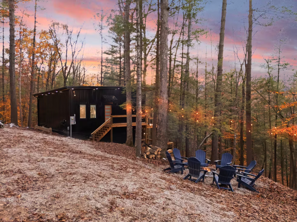 Modern black cabin nestled in the Red River Gorge woods at sunset. Adirondack chairs circle a fire pit, promising a cozy Kentucky getaway.