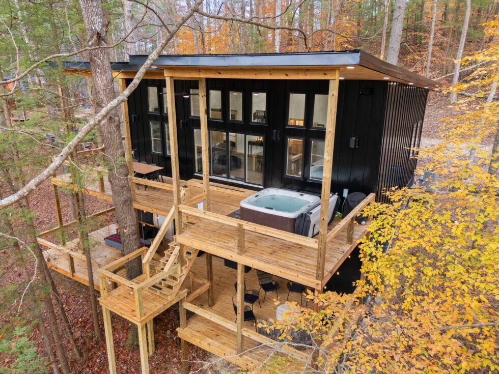 Modern black treehouse cabin rental in Red River Gorge, Kentucky. Elevated decks surround the cabin, one with a hot tub, nestled among autumn trees. Perfect for a unique getaway.