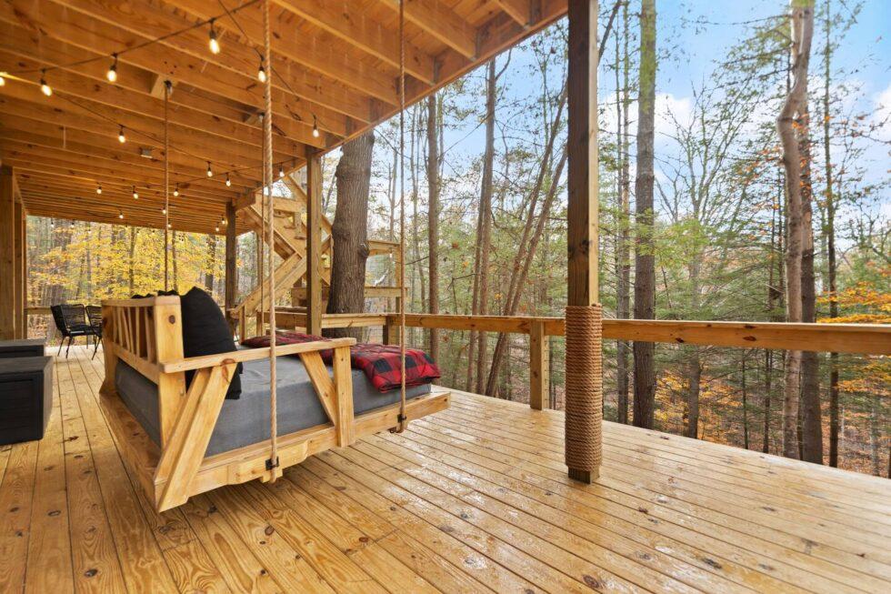Cozy cabin porch swing in Red River Gorge, Kentucky. Wooden swing bed with red plaid blanket hangs beneath string lights, offering a relaxing view of the surrounding forest.